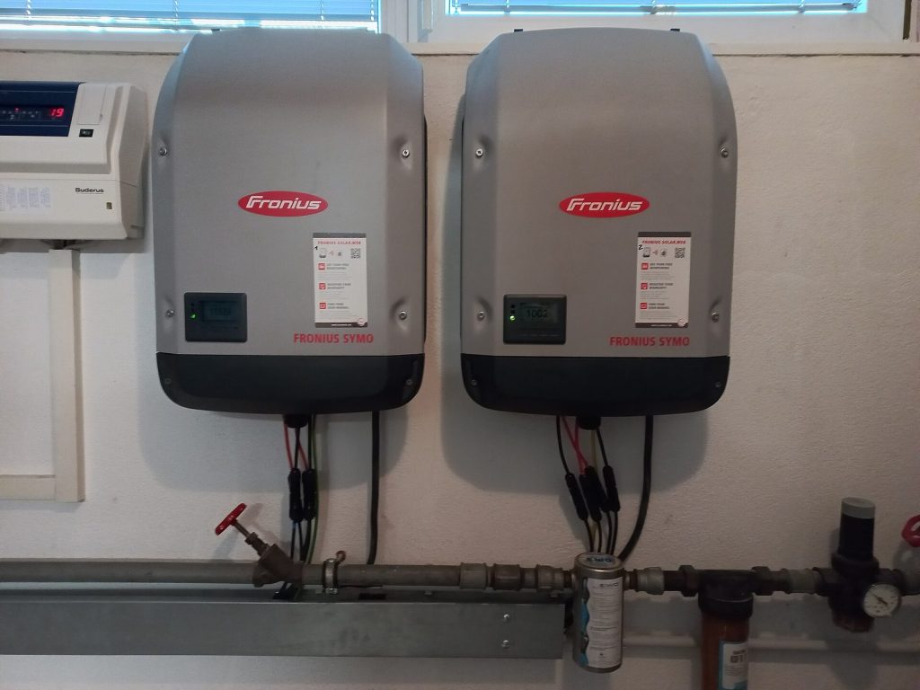 Two Fronius Symo inverters mounted side by side on a utility-room wall above water pipes