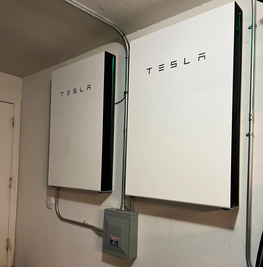 Two white Tesla Powerwall 3 batteries mounted side by side on an indoor wall of a home, with conduit and a grey disconnect box below